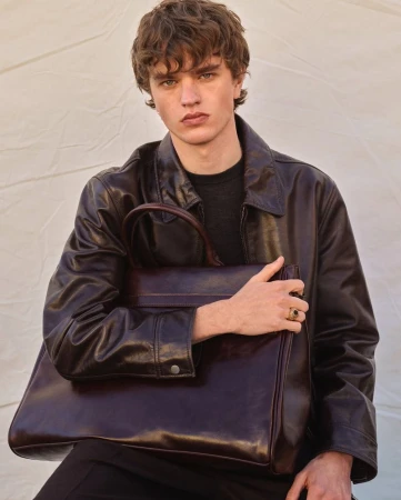 A young male model wearing a dark leather jacket holding a dark leather tote bag against a pale textured background for Zara Origins Spring Summer 2025.