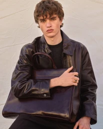A young male model wearing a dark leather jacket holding a dark leather tote bag against a pale textured background for Zara Origins Spring Summer 2025.