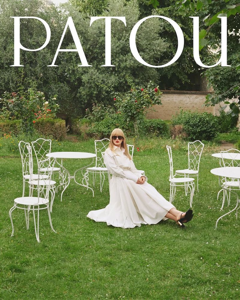 A woman in a white dress poses on a lawn surrounded by metal chairs and tables in a garden setting with the Patou logo overhead.