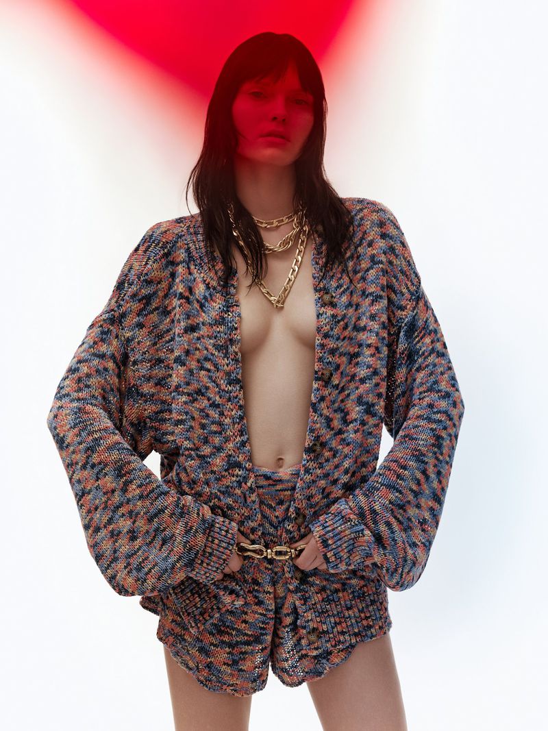 Brunette woman poses in a multicolor knitted cardigan and shorts with gold chain necklace and belt against a white and red background.
