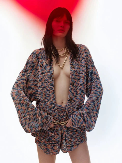 Brunette woman poses in a multicolor knitted cardigan and shorts with gold chain necklace and belt against a white and red background.