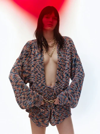 Brunette woman poses in a multicolor knitted cardigan and shorts with gold chain necklace and belt against a white and red background.