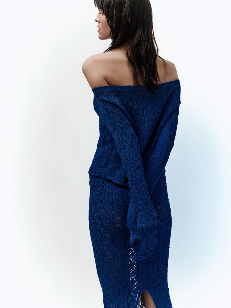 Model seen from behind wearing an off-the-shoulder blue knit top and matching skirt against a white studio background.