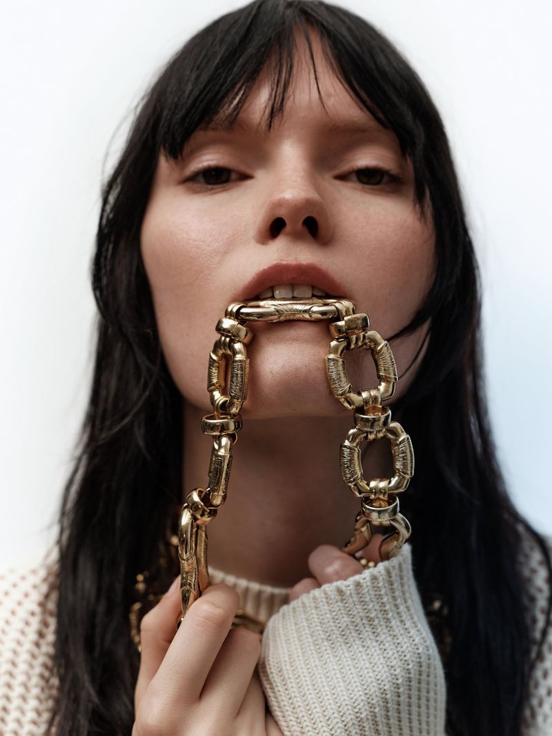 Close-up portrait of a woman with dark hair biting a chunky gold chain necklace, wearing a cream sweater.