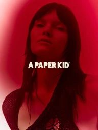 Model with dark hair wearing a crocheted top against a vibrant red gradient background for A Paper Kid SS25.