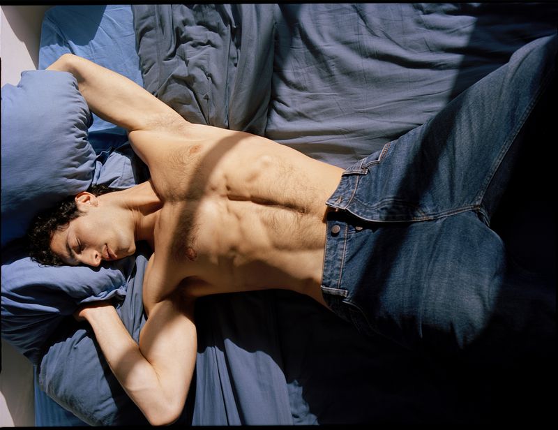 Top-down view of a young adult man with dark hair and jeans lying on a blue bed with sunlight and shadows across his bare torso.
