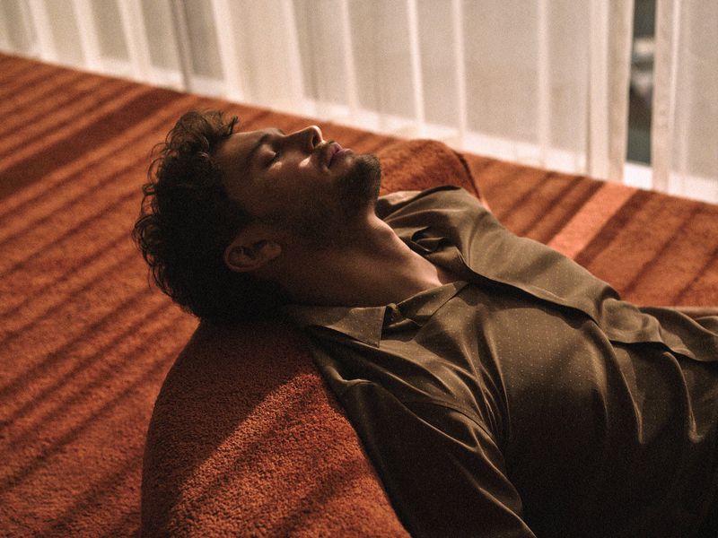 Adult male lies on an orange carpet in natural light wearing a button-down shirt, eyes closed, relaxed. Indoor shot, soft focus.