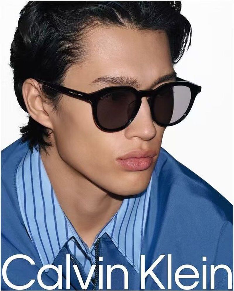 Close-up portrait of a male model wearing black sunglasses and a striped blue shirt against a white backdrop for Calvin Klein