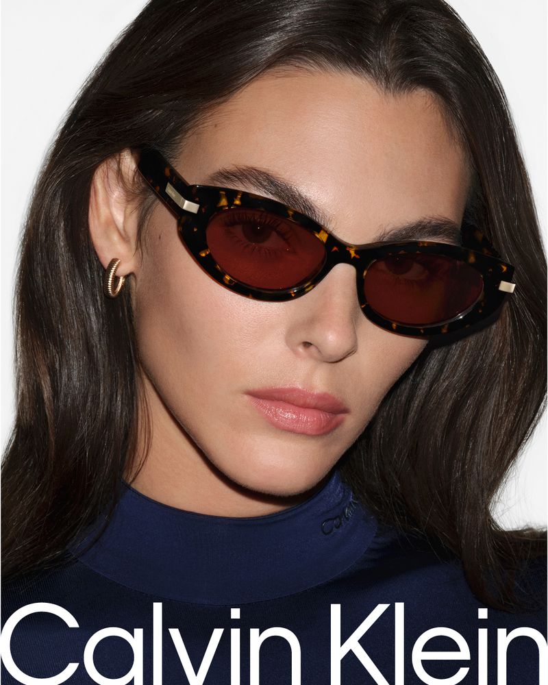 Close-up of a brunette woman wearing tortoise-shell framed sunglasses and a dark-blue Calvin Klein top against a white backdrop.