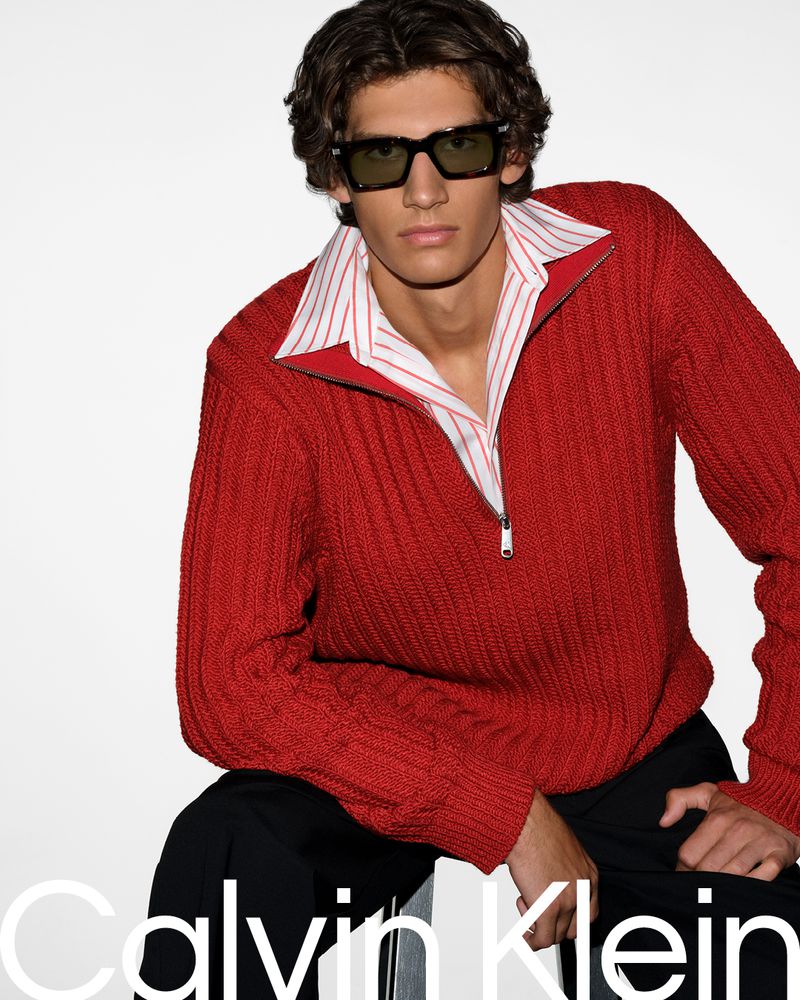 Man in red zip-up sweater and sunglasses poses indoors in a fashion shot for Calvin Klein.