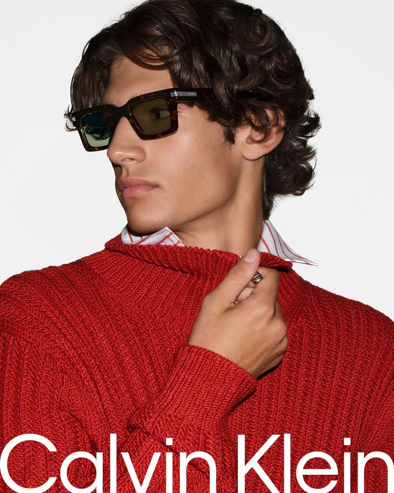 Close-up portrait of a male model wearing tortoiseshell sunglasses and a red ribbed knit sweater against a white background