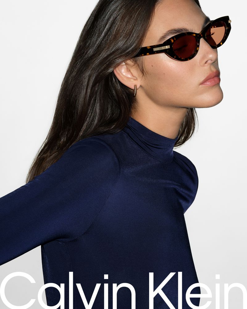 A brunette adult woman in a long-sleeve top poses in a studio wearing tortoise-shell sunglasses with brown lenses, showcasing fashion.