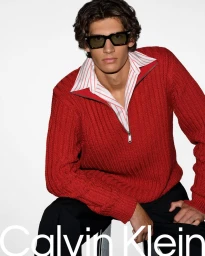 Model wearing dark sunglasses and a red zip-up sweater over a striped shirt against a plain white background