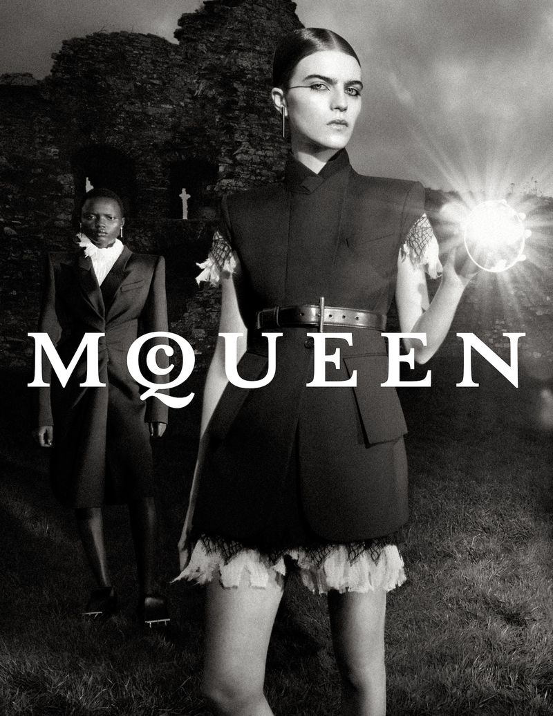 Fashion editorial photo features two models wearing Alexander McQueen designs in a dark outdoor setting with moody monochrome aesthetic.