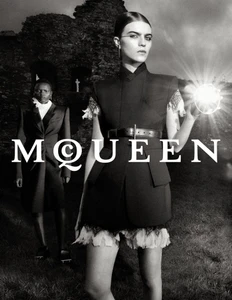 Black and white fashion campaign image featuring two models posing outdoors in structured dark apparel before a ruined stone wall.