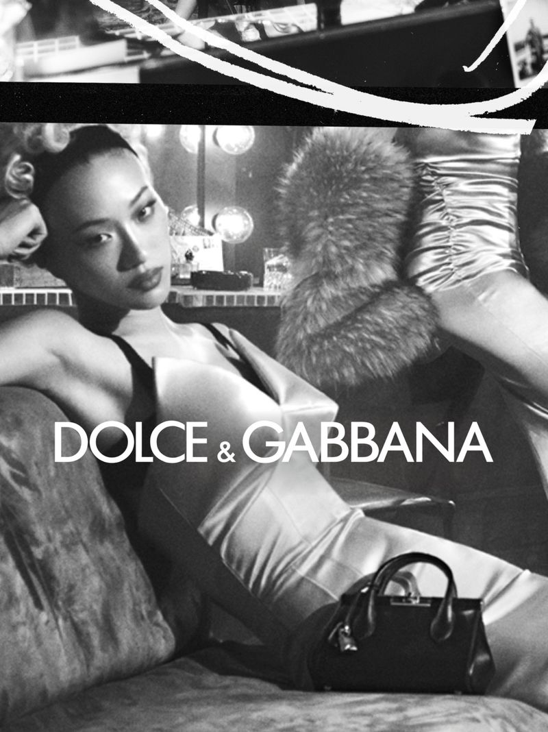 Black and white fashion campaign image featuring a model reclining on a couch wearing a dress, with a handbag in the foreground.