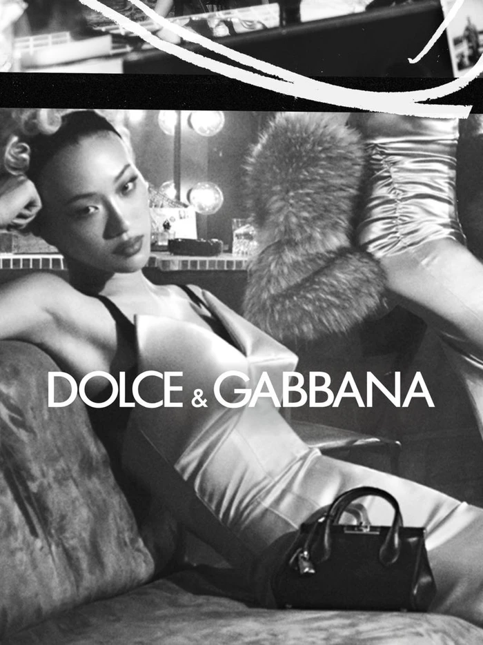 Black and white fashion campaign image featuring a model reclining on a couch wearing a dress, with a handbag in the foreground.