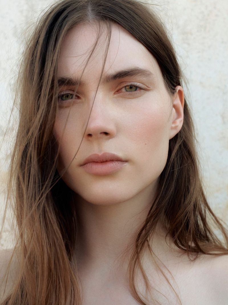 Close-up portrait of a young adult female with long brown hair against a neutral background, emphasizing natural beauty and soft lighting.