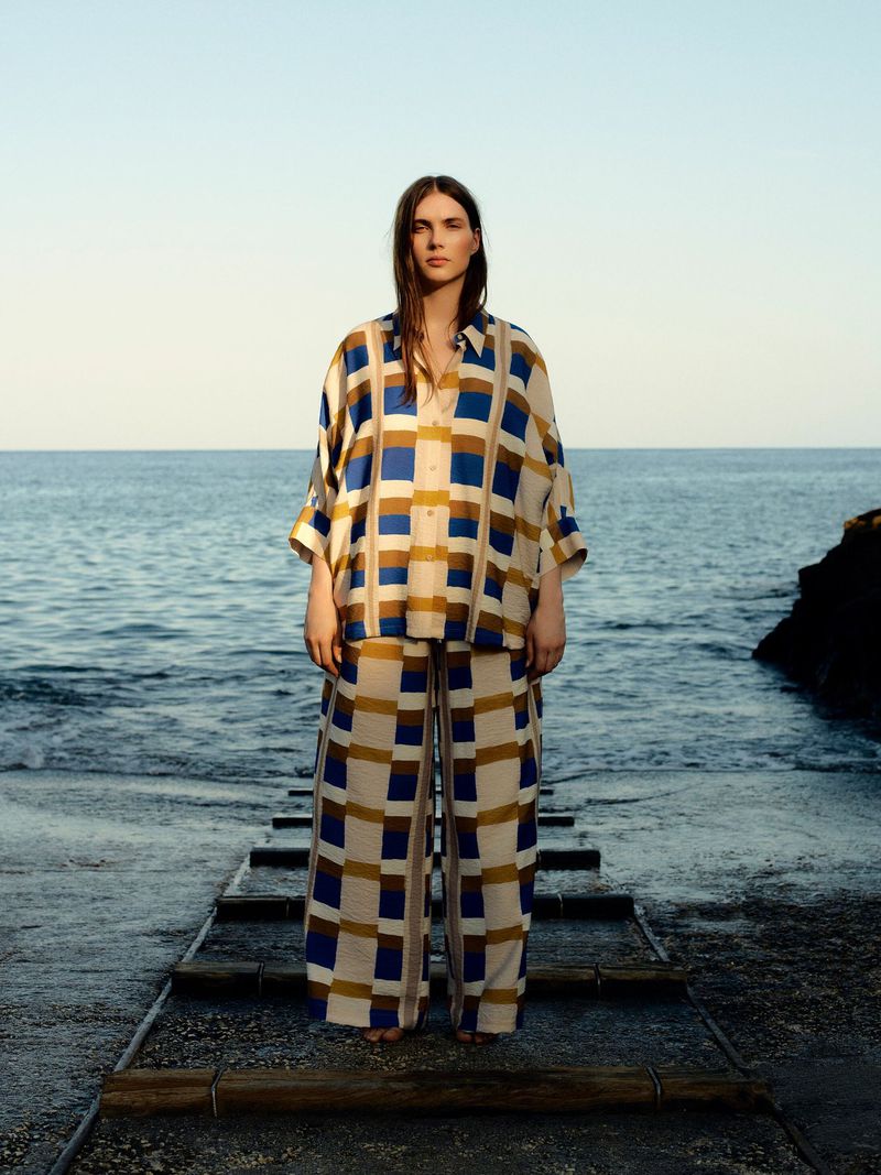 Adult woman stands on weathered steps in matching blue-beige plaid shirt and pants, ocean and clear sky background, fashion editorial.