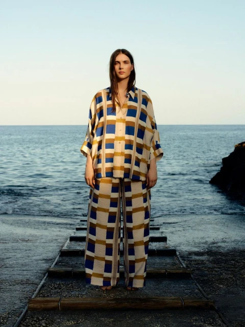 Adult woman stands on weathered steps in matching blue-beige plaid shirt and pants, ocean and clear sky background, fashion editorial.