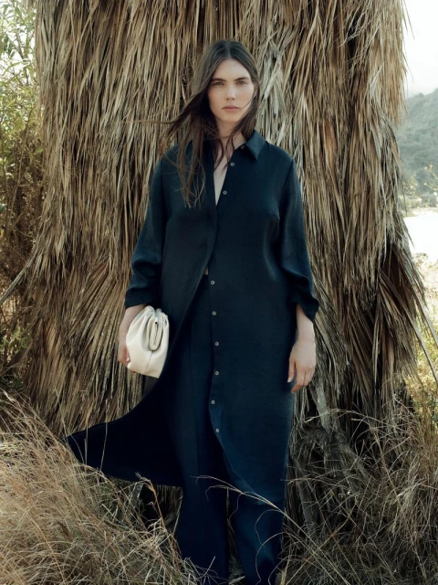 A woman in a dark blue button-down dress holds a cream-colored pouch bag outdoors with a dry palm frond and grassy background.