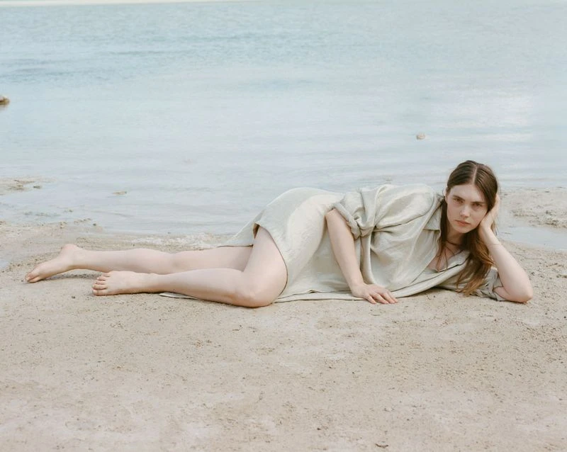 Young woman in a light-colored shirt lays on a sandy beach in a side profile pose with her hand resting on her head near the water.