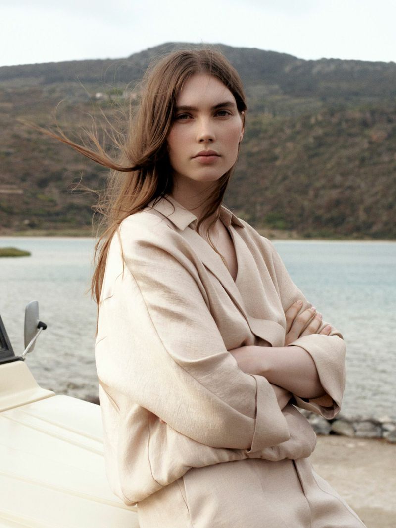 A woman with long brown hair poses in front of a lake and mountains, wearing a beige jacket and pants, with a car in the foreground.