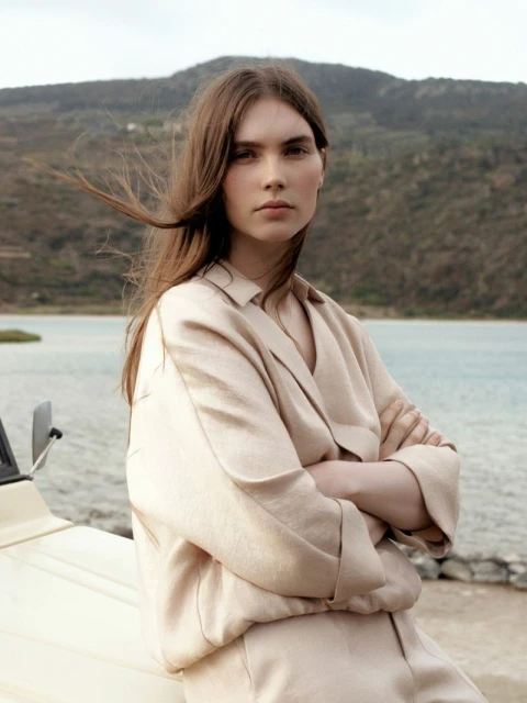A woman with long brown hair poses in front of a lake and mountains, wearing a beige jacket and pants, with a car in the foreground.