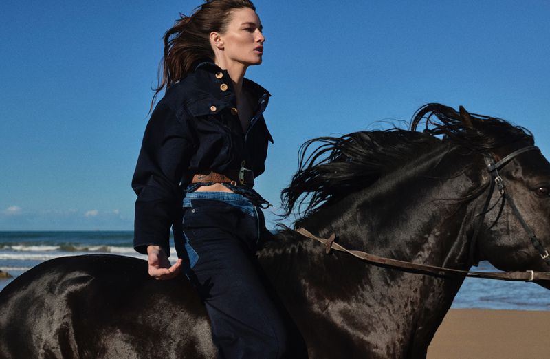 Woman riding a black horse on a beach with blue sky. The model wears denim jacket, jeans and a belt. Sunny day, horse riding lifestyle.