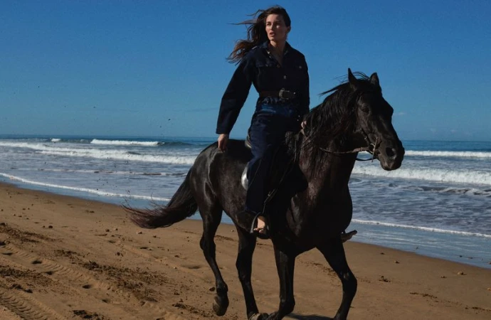 A woman riding a black horse on a sandy beach under a bright blue sky with gentle waves in the background. Equine and human action.