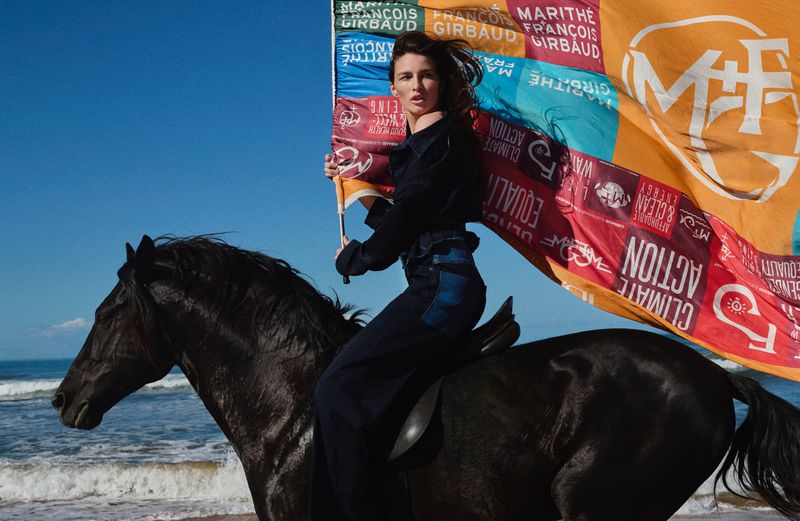 Woman in dark blue attire riding a black horse on a sandy beach, holding a colorful flag with text against a blue sky and ocean.
