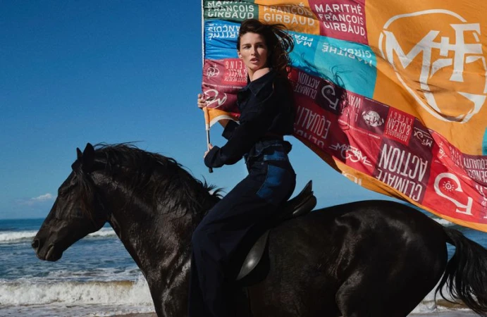 Woman in dark blue attire riding a black horse on a sandy beach, holding a colorful flag with text against a blue sky and ocean.