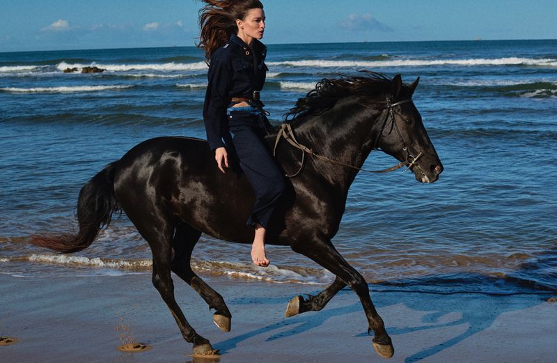 Woman rides a black horse on a beach along the ocean, wearing a dark outfit and no shoes, with the sea and sky in background.