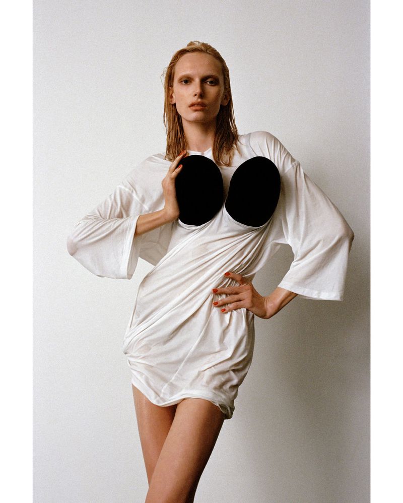 Fashion model poses in a white dress against a neutral backdrop, hands on hips, showcasing edgy style and modern design.