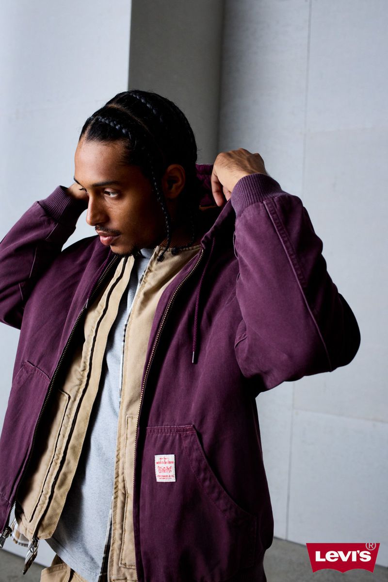 A young man wearing a purple zip-up hoodie poses indoors. The shot features the Levi's logo, with the clothing having a visible tag.