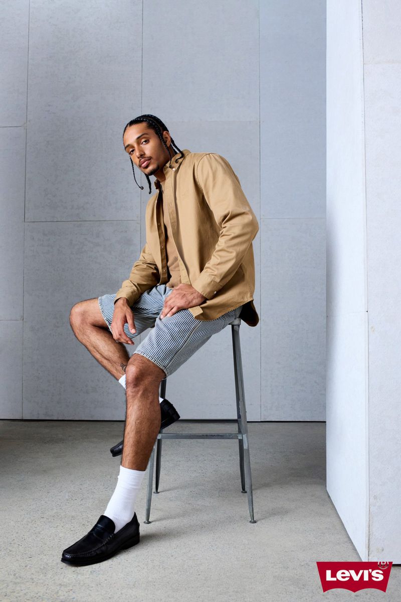 Adult male wearing a tan shirt and striped shorts sits on a stool indoors, wearing black shoes and white socks. Levis logo visible.