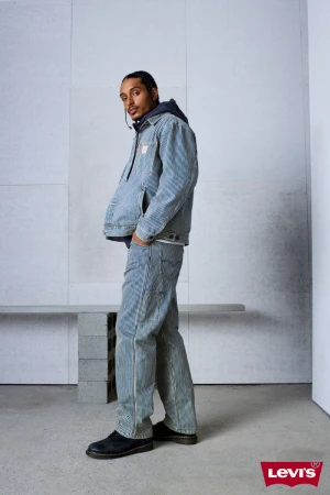 Man wearing matching blue striped denim jacket and trousers standing against a light grey studio wall.