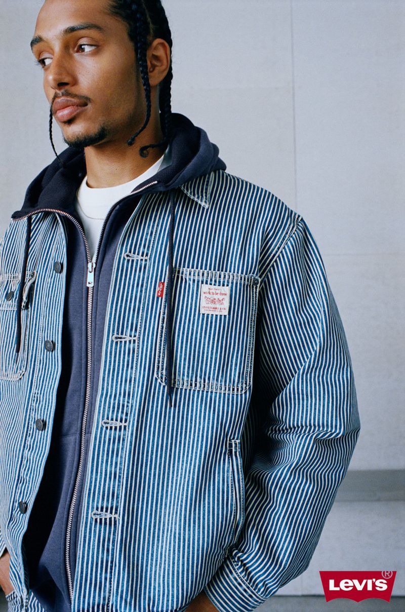 Man wearing a blue striped denim jacket over a dark zip-up hoodie against a light grey wall