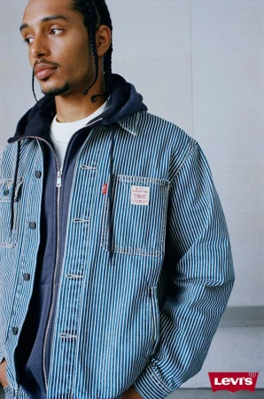 Man wearing a blue striped denim jacket over a dark zip-up hoodie against a light grey wall