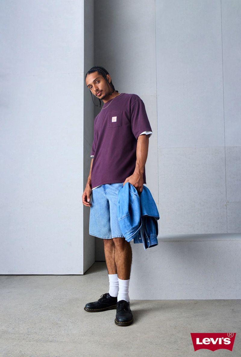 Man in purple t-shirt and jean shorts holds denim jacket against a gray wall. Levi's logo visible in the lower right corner.