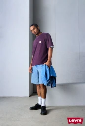 Man wearing a purple t-shirt, denim shorts, and black shoes, holding a denim jacket against a white studio background.