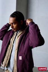Man in a purple hooded zip jacket adjusting his hood against a pale architectural background.