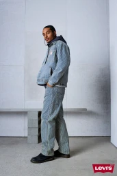 Man wearing matching blue striped denim jacket and trousers standing against a light grey studio wall.