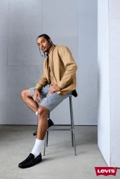 Man seated on a metal stool wearing a tan button-up shirt and patterned shorts against a minimalist grey architectural wall.