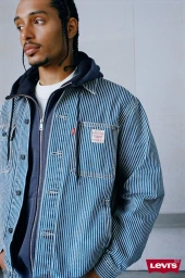 Man wearing a blue striped denim jacket over a dark zip-up hoodie against a light grey wall