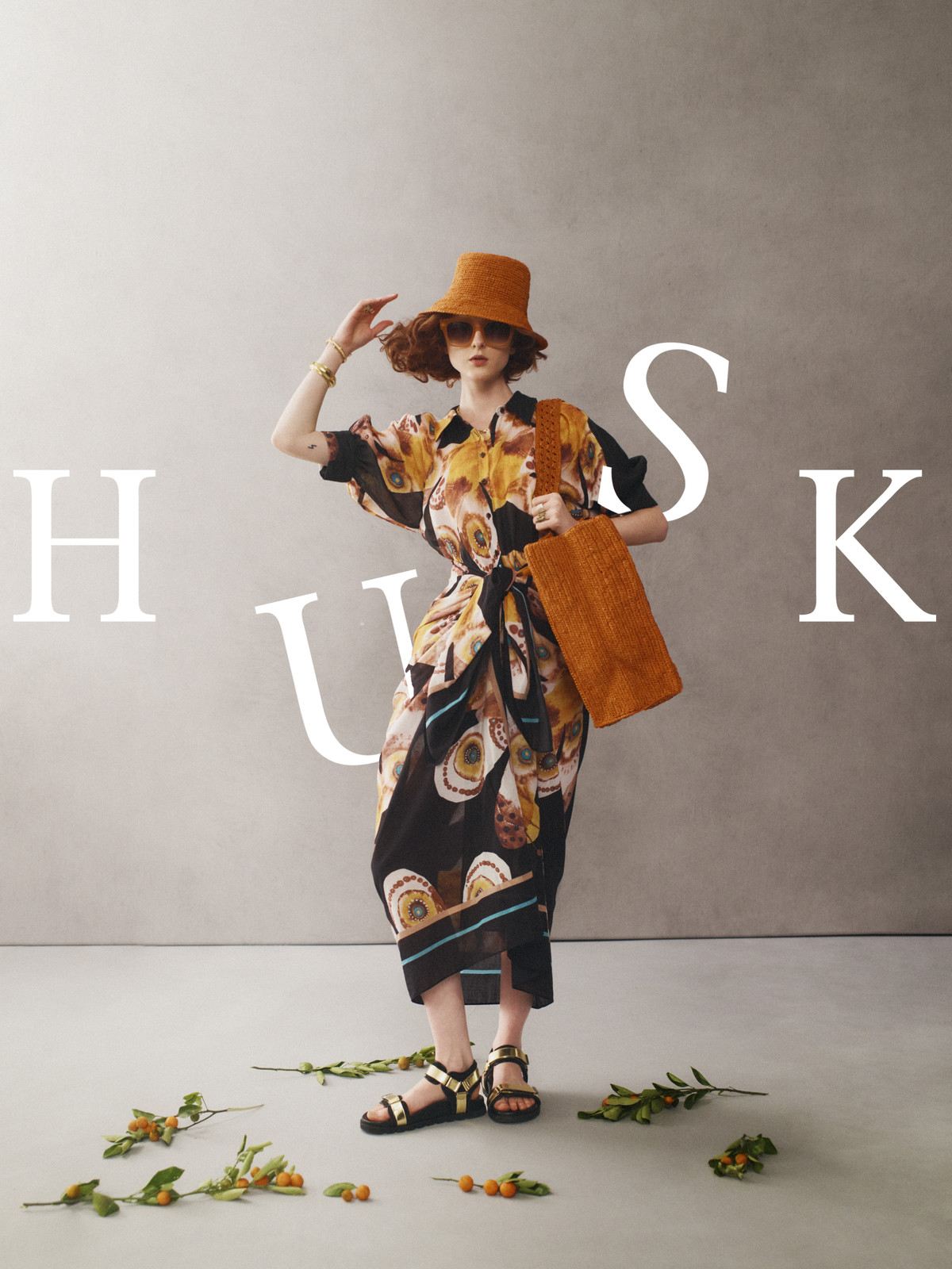 Woman in patterned dress, hat, and sandals stands with bag against neutral backdrop with foliage accent, fashion editorial style.