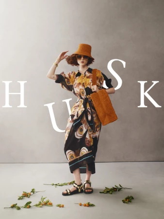 Woman in patterned dress, hat, and sandals stands with bag against neutral backdrop with foliage accent, fashion editorial style.