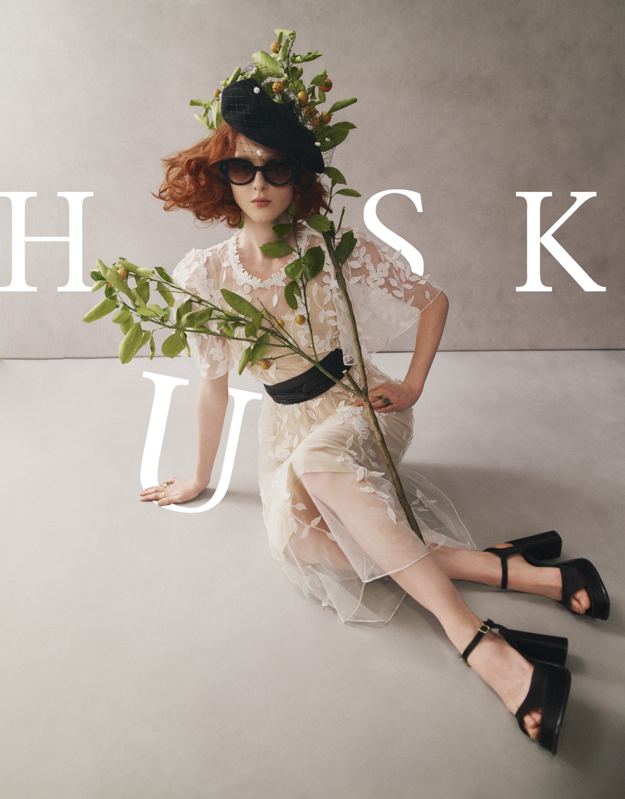 A fashionable redhead poses while seated, wearing a black floral hat, black sunglasses, and sheer dress with black belt.  