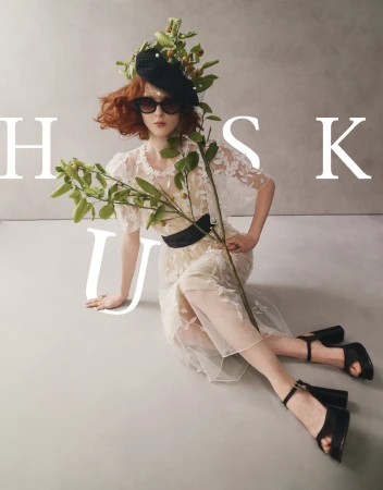 A fashionable redhead poses while seated, wearing a black floral hat, black sunglasses, and sheer dress with black belt.  