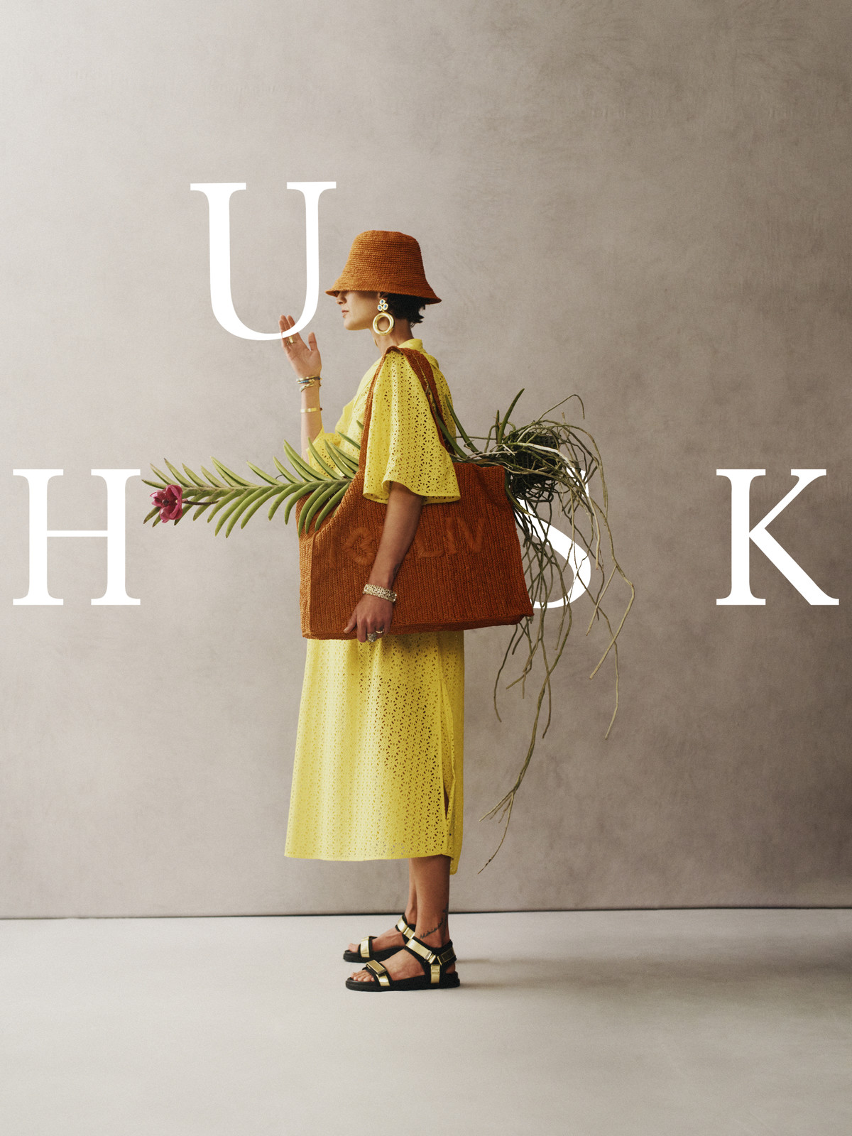 A stylish adult female is posing with a brown tote bag, wearing a yellow dress, straw hat, and sandals against a neutral backdrop.